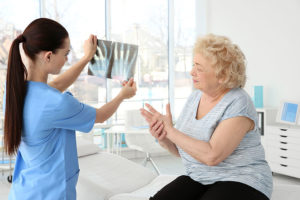 Médica está olhando o raio X das mãos de uma senhora enquanto ela está na cadeira segurando suas mãos com dor por causa da artrose