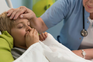 Garoto embaixo das cobertas tossindo o que indica pneumonia infantil. Uma médica passa a mão na cabeça do menino.