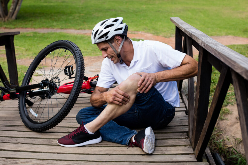 Homem mais velho sentado no chão de uma ponte com as mãos no joelho por causa de uma lesão provocada pelo ciclismo. Ele está de capacete e a bicicleta está logo atrás dele.