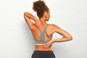 Mulher de costas com as mãos na região superior das costas, do pescoço, dos ombros e da lombar indicando que ela está com dores provenientes da fibromialgia. Essas regiões estão marcadas de vermelho.