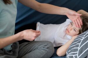 Menina com pneumonia infantil na cama com a mãe medindo a sua temperatura e colocando a mão na sua testa