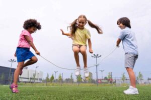 Três crianças pulando corda ao ar livre. Duas meninas e 1 menino. Representação da importância da atividade física na infância.