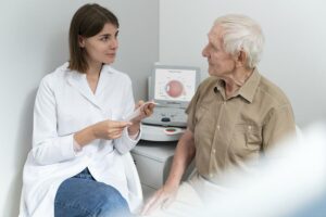 Oftalmologista mulher atendendo paciente idoso. Eles estão se olhando e ela está com um papel do resultado do exame. Atrás deles tem o aparelho do exame com a anatomia de um olho na tela. Esse senhor está com suspeita de glaucoma, por isso foi se consultar.