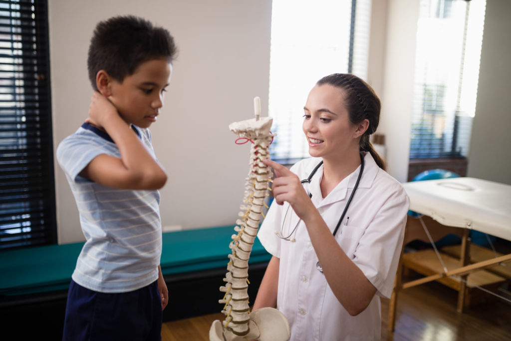 Garoto com a mão no pescoço indicando torcicolo olhando para um médica que está com um modelo de uma coluna apontando para as vertebras da região do pescoço