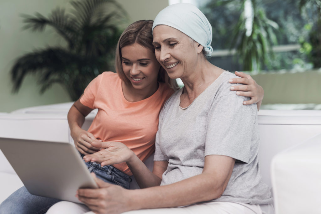 Duas mulheres, uma saudável e outra com câncer, sentadas e abraçadas em um sofá olhando sorridentes para um tablet