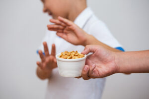 Close de uma mão com um pote de amendoim enquanto, ao fundo, um garoto desfocado rejeita o alimento por ter alergia alimentar