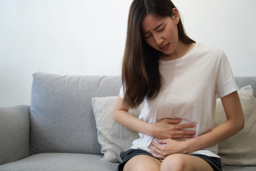 Mulher asiática com sentada no sofá com as mão na barriga e o semblante de dor por causa da síndrome do intestino irritável
