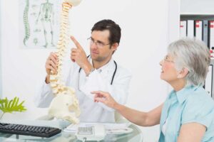 Médico ortopedista mostrando um modelo de coluna para uma paciente idosa. Representação de como funciona uma consulta com o ortopedista.