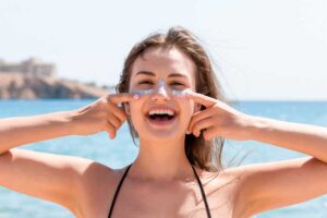 Mulher sorridente aplicando protetor solar em seu rosto. Ao fundo, o mar. Representação dos cuidados com a pele no verão.