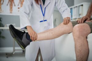 Médica ortopedista apalpando a perna de um paciente idoso. O paciente está sentado na cama do consultório e a médica ortopedista está apalpando o joelho e a canela, Representação dos cuidados ortopédicos na terceira idade.